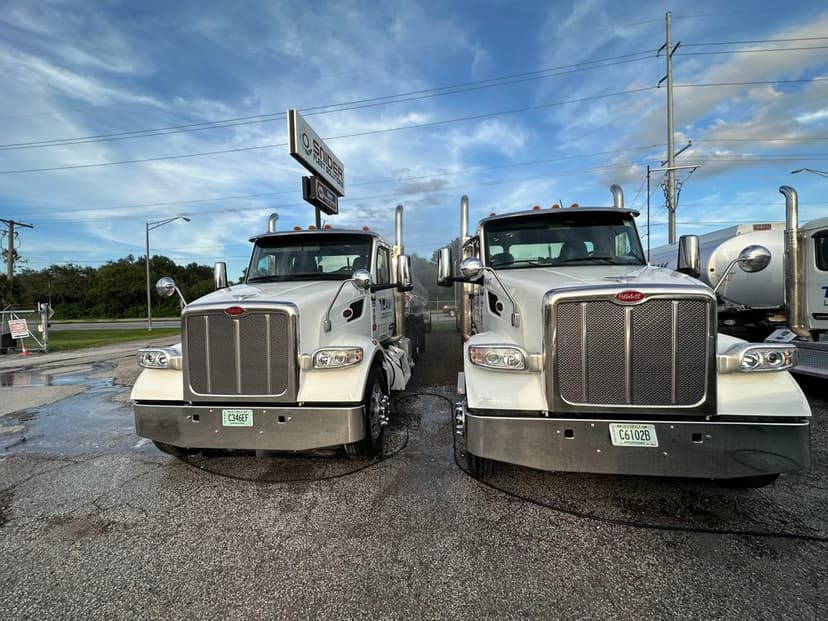 Two silver Peterbilt trucks being washed at a truck wash under a blue sky.